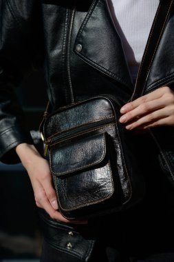 Siyah deri ceketli şık bir manken eşsiz siyah deri bir çanta, parlak bir parıltı ve ön cepli bir çanta sergiliyor.