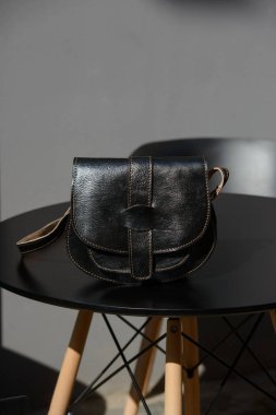 Benzersiz bir kapağı ve zıt dikişleri olan küçük, siyah deri bir çapraz-vücut torbası, güneşli bir açık hava ortamında modern bir masada sergileniyor..