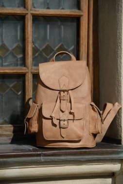 Ayrıntılı tokaları ve cepleri olan yüksek kaliteli bej bir sırt çantası kırsal bir pencere pervazında sunuluyor. Zarif, klasik tasarımı vurgulanıyor.
