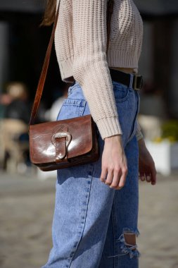 Örgü kazak ve yırtık kot giyen şık bir manken şehir sokaklarında belirgin bir kemer tokası olan şık kahverengi deri bir çanta sergiliyor.