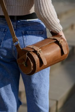 Örgü süveter ve kot pantolon giyen bir mankenin yakın plan fotoğrafı. Eşsiz bir silindirik deri çanta, karmaşık bir yan detay ve kemer takılı..