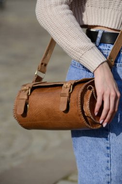 Örgü süveter ve kot pantolon giyen bir mankenin yakın plan fotoğrafı. Eşsiz bir silindirik deri çanta, karmaşık bir yan detay ve kemer takılı..