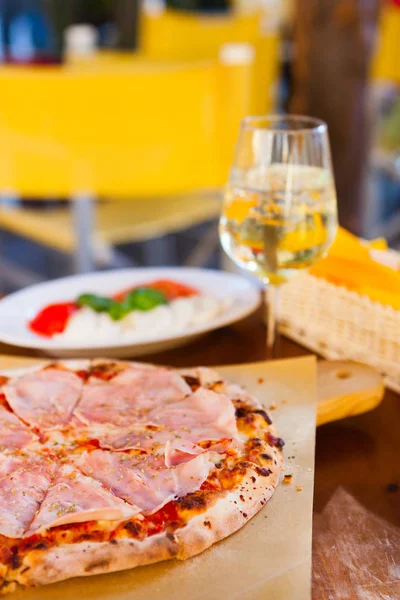 Italian food dinner. Pizza with tomato prosciutto mozzarella and glass white wine summer outdoor restaurant, selective focus