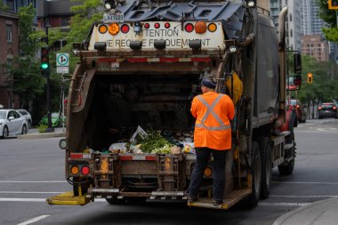 Kanada, 06 Temmuz 2025: Çöp toplayıcısı turuncu yansıtıcı güvenlik ceketi giyerek kamyonun arkasına biniyor