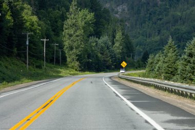 Quebec, Kanada, 20 Temmuz 2025: Ormanlık arazi, araba ve sarı uyarı işareti ile kırsal otoyolun kıvrımı