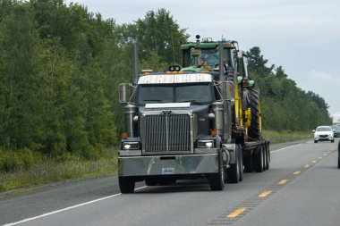 Kanada, 20 Temmuz 2025: Büyük siyah kamyonet, yeşil traktörü kırsal ağaçlarla kaplı otoyoldan aşağı çekiyor