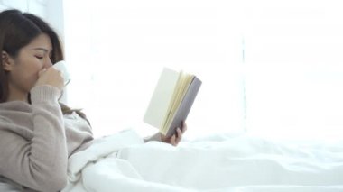 Güzel Asyalı kadın sıcak kahve içme ve yatak odasında yatakta kitap okuma zevk. Bir kitap ve coffee.lifestyle Asya kadın fincan ev concept tutarak rahat kazak giyen Asya erkek