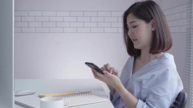 Evde oturma odasında otururken dizüstü bilgisayarda çalışma güzel genç gülümseyen Asyalı kadın. Ev ofisinde çalışmak için telefon kullanarak Asya iş kadını. Evde zaman zevk.