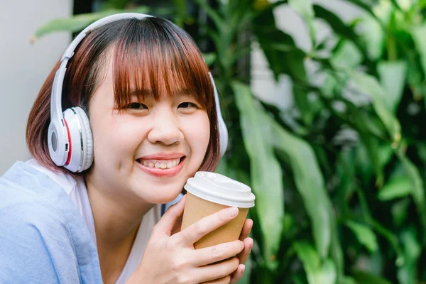 Happy young Asian woman listening to music in a coffee shop holding a cup of coffee in her hand. Young Woman listening to music with headphone while relaxing in the garden. Coffee relax concept.