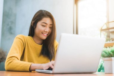 Onun gülümseme ile masa üzerinde laptop ile çalışan genç Asyalı kadın. Onu laptop ile hafta sonu sıcak güneş ışığı gün çalışan genç kadın. Dizüstü ev kavramı içinde çalışma.