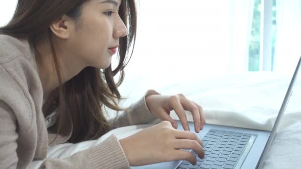 Krásná mladá usměvavá Asijské žena pracuje na notebooku, zatímco sedí na posteli v ložnici doma. Asijské ženy relaxovat ve svém domě. Využívat čas doma.