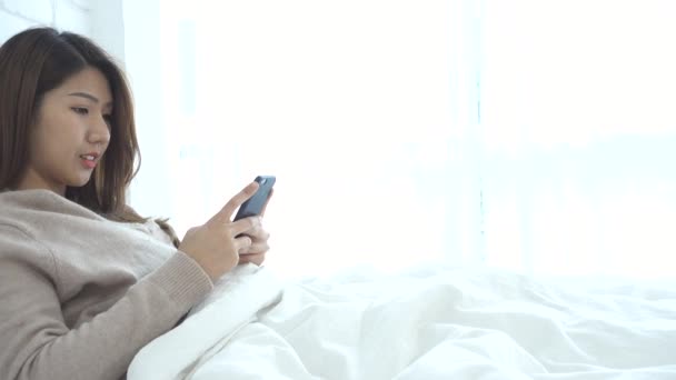 幸せなアジア女性は ベッドの上のスマート フォンを使用しています スマート フォン ソーシャルアプリをチェック ベッドでアジアの女性 笑顔の女性は 家庭での携帯電話でネット サーフィンします — ストック動画