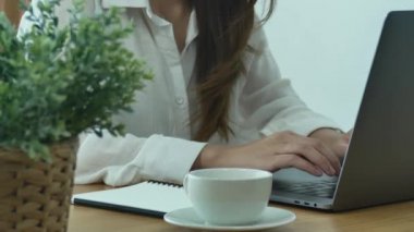 Bir evde oturma odasında sıcak kahve içme tadını çıkarırken dizüstü bilgisayarda çalışan güzel genç gülümseyen kadın. Evde zaman zevk. Ev ofisinde çalışan Asya iş kadın.