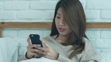 Mutlu Asyalı kadınlar akıllı telefon sabah yatakta kullanıyorsunuz. Asyalı kadın yatakta smartphone ile sosyal uygulamalar kontrol. Gülümseyen kadın evde cep telefonu ile net sörf. Telefon bağımlısı kavramı.