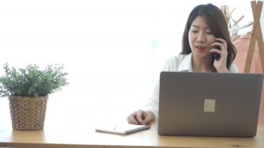 Evde oturma odasında otururken dizüstü bilgisayarda çalışma güzel genç gülümseyen Asyalı kadın. Ev ofisinde çalışmak için telefon kullanarak Asya iş kadını. Evde zaman zevk.