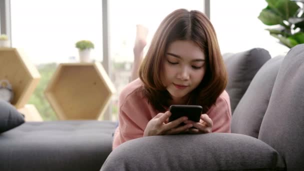 亚洲女人躺在客厅的沙发上使用智能手机 快乐的女性在家里用电话发短信 发短信和上网购物 家庭妇女的生活方式概念 — 图库视频影像