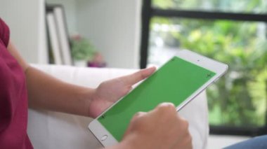 Genç Asyalı kadın ile yeşil ekran siyah tablet cihaz kullanarak. Oturma odasında kanepede otururken sayfalarını kaydırma Asyalı kadın tablet, holding. Chroma anahtar.