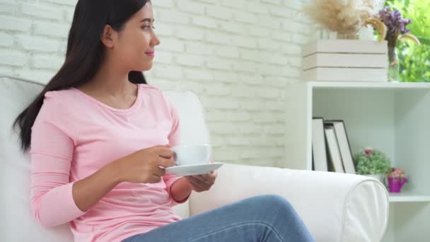 快乐的亚洲年轻女子 她在家里的起居室里喝着温暖的咖啡或茶 享受着它 有魅力的亚洲女人捧着一杯咖啡 — 图库视频影像
