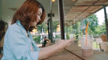 Neşeli mutlu Asyalı Genç kadın oturma içme buzlu yeşil çay konuşma, okuma ve manifatura için smartphone kullanarak kafede. Kadın yaşam tarzı kavramı.