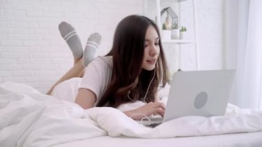 Güzel Asyalı kadın yatak odasında yatağın üstüne yalan sırasında bilgisayar oynarken. Mutlu kadın dinleme müzik arama şarkıları evde bir laptop ve kulaklık ile. Yaşam tarzı kadına ev concept.