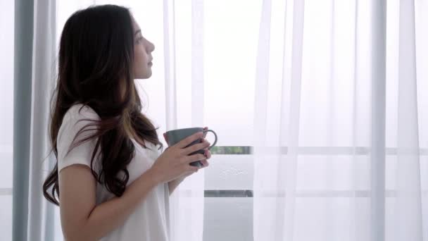 年轻的亚洲妇女在她家卧室的湿窗旁享受咖啡或茶 美丽的青少年女性饮用饮料和在雨天放松 — 图库视频影像