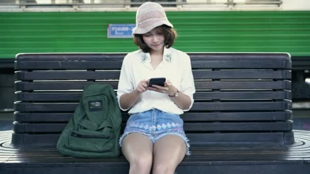 旅行者背包亚洲妇女在泰国曼谷旅游 坐在长凳上的快乐年轻女性使用智能手机在火车站交谈 阅读和发短信 — 图库视频影像