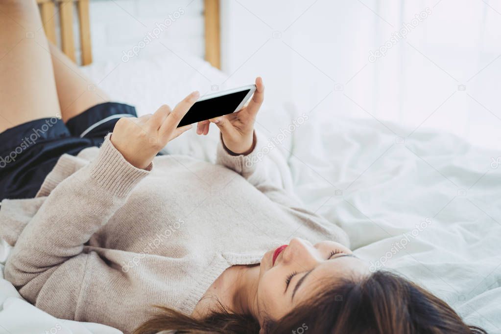 Happy Asian women are using smart phone with black blank empty screen on the bed in morning. Asian woman in bed checking social apps with smartphone. Smiling woman surfing net with cellphone at home.