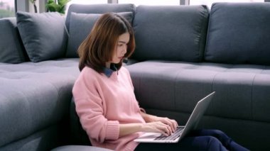 Evde oturma odasında kahve içme tadını çıkarırken dizüstü bilgisayarda çalışma güzel genç gülümseyen Asyalı kadın. Mutlu kadın evde alışveriş online satın alma. Yaşam tarzı kadına ev concept.