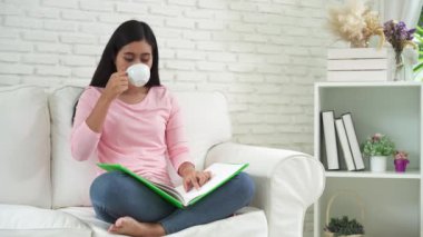 İlginç bir kitap ve içme sıcak çay veya kahve beyaz Kupası evde rahat oturma odasında kanepede okuma Genç Asyalı çekici kadın. Rahat yaşam tarzı evde.
