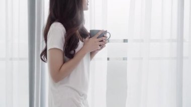 Gülümseyen ve yatak odasında pencere yakınındaki bir fincan kahve ya da çay içme mutlu güzel Asyalı kadın. Genç Asya kadın Perdeleri açın ve sabah sakin ol. Yaşam tarzı kadına ev concept.