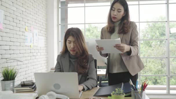 Attractive smart Asian business woman in smart casual wear working on laptop while sitting on desk while her angry manager standing angry throwing papers on office desk. Women work at office concept. 