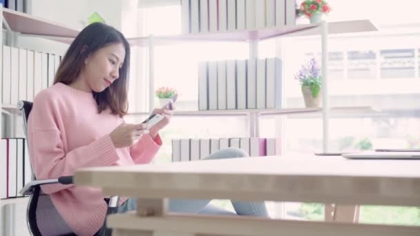 美丽的亚洲妇女使用智能手机购买信用卡网上购物 而穿毛衣坐在办公桌上的客厅在家里 生活方式妇女在家的概念 — 图库视频影像