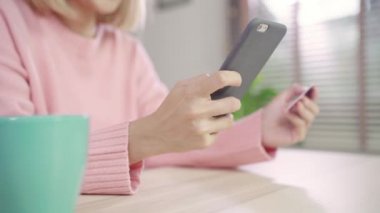 Güzel Asyalı kadın Smartphone Resepsiyon evde oturma odasında oturup giyim kazak sırasında kredi kartıyla alışveriş online satın alma kullanarak. Yaşam tarzı kadına ev concept.