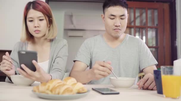 有吸引力的年轻亚裔夫妇在餐桌上用报纸和手机分心 同时吃早餐 兴奋的年轻亚裔夫妇惊讶于令人难以置信的好消息 快乐的家庭惊讶互联网 — 图库视频影像