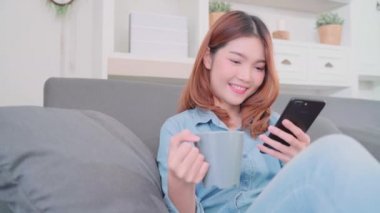 Güzel çekici gülümseyen Asyalı kadın sıcak bir fincan kahve ya da çay koltukta yalan Ise tutarak smartphone kullanarak portresi ne zaman evde oturma odasında gevşeyin. Yaşam tarzı kadınlara ev concept.