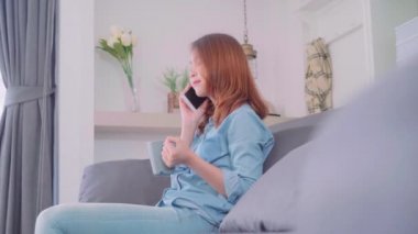 Güzel çekici gülümseyen Asyalı kadın sıcak bir fincan kahve ya da çay koltukta yalan Ise tutarak smartphone kullanarak portresi ne zaman evde oturma odasında gevşeyin. Yaşam tarzı kadınlara ev concept.