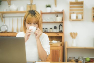 Bir evde oturma odasında sıcak kahve içme tadını çıkarırken dizüstü bilgisayarda çalışan güzel genç gülümseyen kadın. Evde zaman zevk. Ev ofisinde çalışan Asya iş kadın.