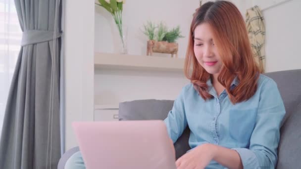美しい魅力的な若い笑顔アジアの女性の肖像 ソファの上に横たわっている間コンピューターまたはラップトップを使用して 自宅のリビング ルームでリラックスします ホーム コンセプトでライフ スタイルの女性を時間を楽しんでください — ストック動画