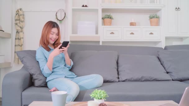 美丽迷人的年轻微笑的亚洲妇女的肖像使用智能手机 而躺在沙发上放松在客厅在家里 享受时间生活方式的妇女在家里的概念 — 图库视频影像