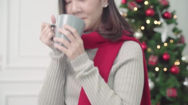 陽気な幸せな若いアジア女性コーヒー クリスマス フェスティバルで彼女のリビング ルームを自宅でカメラに笑顔の暖かいカップを保持しています ライフ スタイルの女性を祝うクリスマスと新年のコンセプト — ストック動画