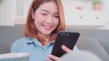 Güzel çekici gülümseyen Asyalı kadın sıcak bir fincan kahve ya da çay koltukta yalan Ise tutarak smartphone kullanarak portresi ne zaman evde oturma odasında gevşeyin. Yaşam tarzı kadınlara ev concept.