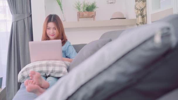 美しい魅力的な若い笑顔アジアの女性の肖像 ソファの上に横たわっている間コンピューターまたはラップトップを使用して 自宅のリビング ルームでリラックスします ホーム コンセプトでライフ スタイルの女性を時間を楽しんでください — ストック動画
