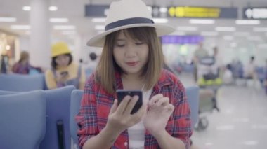 Kullanarak ve onun smartphone uçağının kalkış kapısı uluslararası havaalanında beklerken terminal Salonu sandalyede oturan süre kontrol mutlu Asyalı kadın. Kadınlar Havaalanı kavramında mutlu.