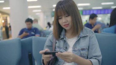 Kullanarak ve onun smartphone uçağının kalkış kapısı uluslararası havaalanında beklerken terminal Salonu sandalyede oturan süre kontrol mutlu Asyalı kadın. Kadınlar Havaalanı kavramında mutlu.