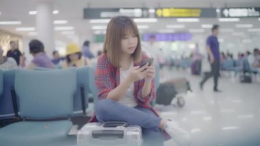 Kullanarak ve onun smartphone uçağının kalkış kapısı uluslararası havaalanında beklerken terminal Salonu sandalyede oturan süre kontrol mutlu Asyalı kadın. Kadınlar Havaalanı kavramında mutlu.