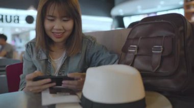 Kullanarak ve onun smartphone uçağının kalkış kapısı uluslararası havaalanında beklerken terminal Salonu sandalyede oturan süre kontrol mutlu Asyalı kadın. Kadınlar Havaalanı kavramında mutlu.