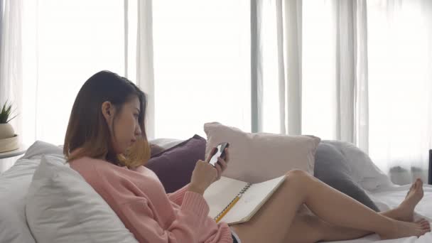 幸せな若いアジア女性自宅で勉強は自宅のソファで横になっている笑顔で彼女の携帯電話に Sms またはテキスト メッセージを読む — ストック動画
