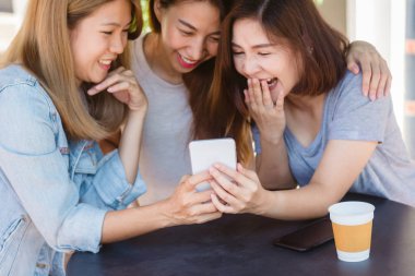 Arkadaş grubu kahve içme ve birlikte konuşurken kafede oturan neşeli Asyalı Genç kadınlar. Kahve için konuşma, okuma ve manifatura smartphone kullanırken zevk çekici Asyalı kadın.