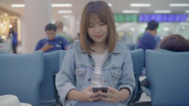 Kullanarak ve onun smartphone uçağının kalkış kapısı uluslararası havaalanında beklerken terminal Salonu sandalyede oturan süre kontrol mutlu Asyalı kadın. Kadınlar Havaalanı kavramında mutlu.