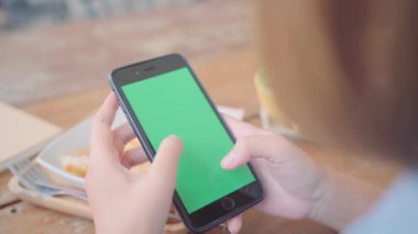 Genç Asyalı kadın ile yeşil ekran siyah cep telefonu cihaz kullanarak. Kafede otururken sayfalarını kaydırma Asyalı kadın smartphone, holding. Chroma anahtar.